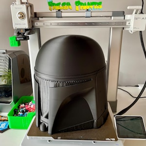 May include: A black Mandalorian helmet is being 3D printed on a Bambu Lab A1 printer. The printer is white and gray, with a green and yellow sign that reads "The Fresh Prints". The helmet is on a gray printing bed.