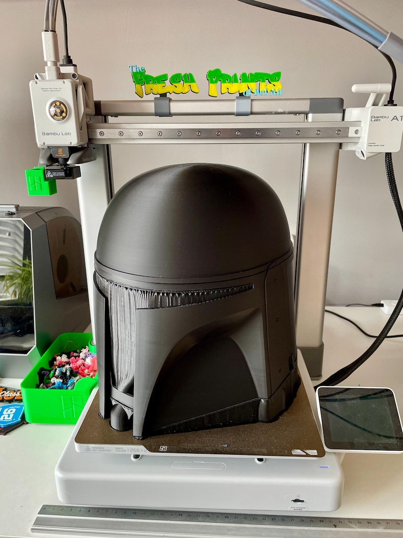 May include: A black Mandalorian helmet is being 3D printed on a Bambu Lab A1 printer. The printer is white and gray, with a green and yellow sign that reads "The Fresh Prints". The helmet is on a gray printing bed.
