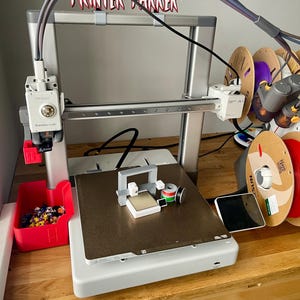 Pu&ograve; includere: Una stampante 3D con telaio argentato e contenitore rosso per i pezzi. La stampante ha un piano di stampa con un piccolo oggetto stampato e diverse bobine di filamento di vari colori. Il testo "Printer Runner" &egrave; visibile sopra la stampante.