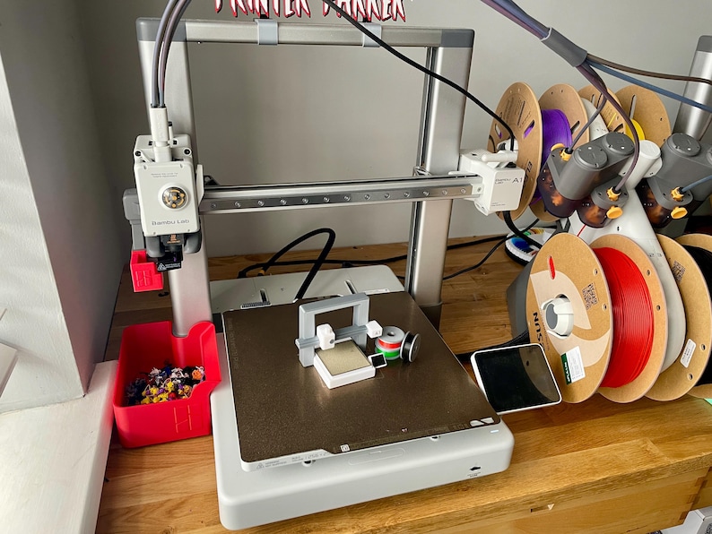 Pu&ograve; includere: Una stampante 3D con telaio argentato e contenitore rosso per i pezzi. La stampante ha un piano di stampa con un piccolo oggetto stampato e diverse bobine di filamento di vari colori. Il testo "Printer Runner" &egrave; visibile sopra la stampante.