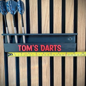 May include: A black dart holder with the text "TOM'S DARTS" in red, holding three darts. The darts have black shafts and blue and silver flights. A measuring tape shows the holder is approximately 10 inches long. The background is a wooden wall.