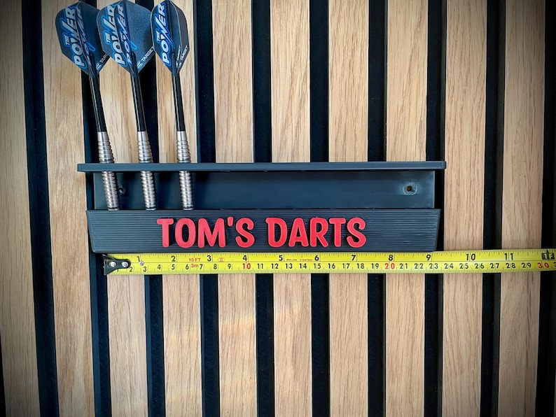 May include: A black dart holder with the text "TOM'S DARTS" in red, holding three darts. The darts have black shafts and blue and silver flights. A measuring tape shows the holder is approximately 10 inches long. The background is a wooden wall.