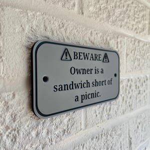 May include: A rectangular, gray metal sign with black trim is mounted on a white brick wall. The sign reads "BEWARE Owner is a sandwich short of a picnic." The sign has two small holes for mounting and a warning symbol.