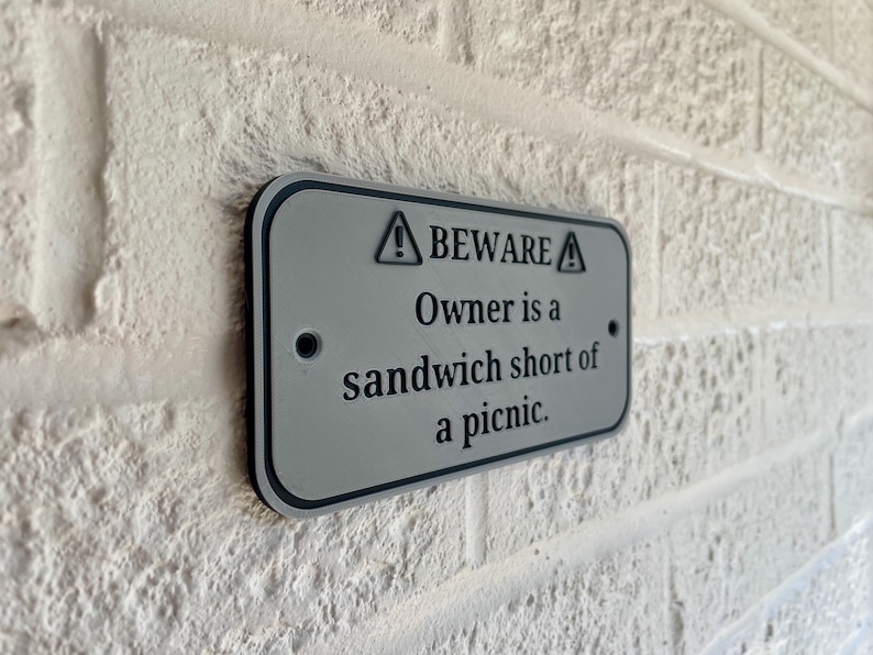 May include: A rectangular, gray metal sign with black trim is mounted on a white brick wall. The sign reads "BEWARE Owner is a sandwich short of a picnic." The sign has two small holes for mounting and a warning symbol.