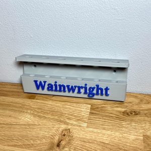 Wall mounted dart holder. Silver with Blue personalised text on the front reading Wainwright