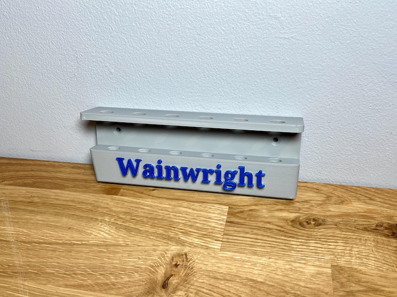 Wall mounted dart holder. Silver with Blue personalised text on the front reading Wainwright