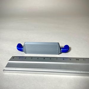 May include: A gray and blue intercooler model with a ruler for scale. The model has a rectangular body with horizontal lines and blue curved pipes on each end. The ruler shows measurements in inches.