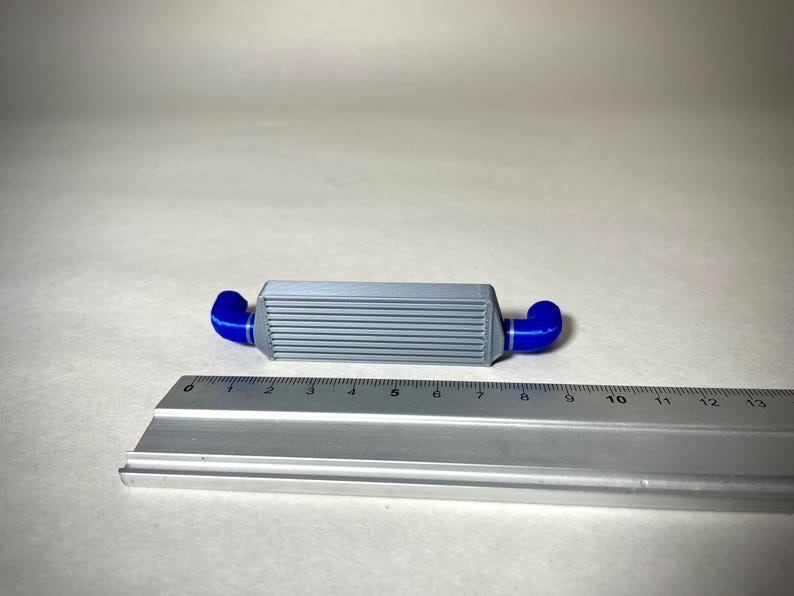 May include: A gray and blue intercooler model with a ruler for scale. The model has a rectangular body with horizontal lines and blue curved pipes on each end. The ruler shows measurements in inches.