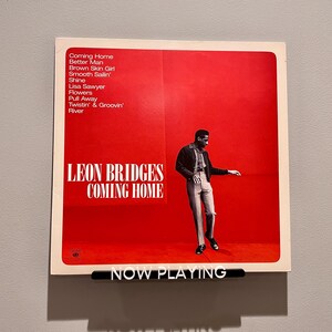 May include: A red album cover with a white border featuring a black and white photo of a man in a jacket and pants. The album cover has the text "LEON BRIDGES COMING HOME" in white letters. The text "NOW PLAYING" is on a black bar at the bottom of the image.