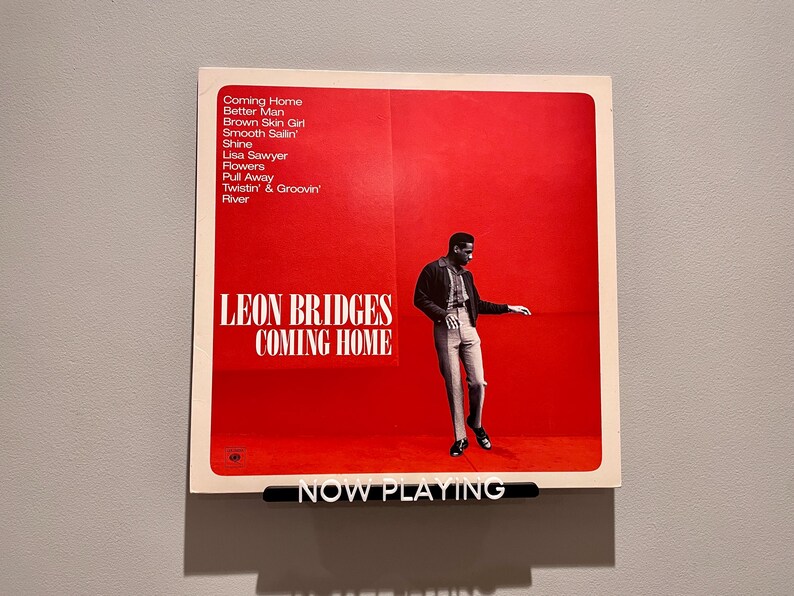 May include: A red album cover with a white border featuring a black and white photo of a man in a jacket and pants. The album cover has the text "LEON BRIDGES COMING HOME" in white letters. The text "NOW PLAYING" is on a black bar at the bottom of the image.