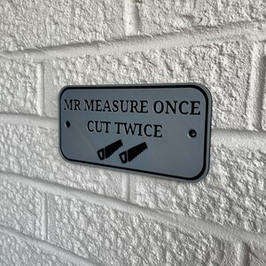 May include: A rectangular gray metal sign with the text "MR MEASURE ONCE CUT TWICE" and two saw illustrations. The sign is mounted on a white brick wall.