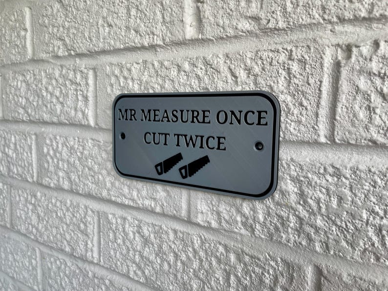 May include: A rectangular gray metal sign with the text "MR MEASURE ONCE CUT TWICE" and two saw illustrations. The sign is mounted on a white brick wall.