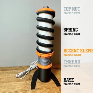 Op de afbeelding: Een zwart-witte veer met oranje accenten. De veer is bevestigd aan een zwarte basis met een zilveren moer. De tekst "TOP NUT EXAMPLE SILVER", "SPRING EXAMPLE BLACK", "ACCENT ELEMENTS EXAMPLE ORANGE", "THREAD EXAMPLE SILVER" en "BASE EXAMPLE BLACK" is zichtbaar aan de rechterkant van de afbeelding.