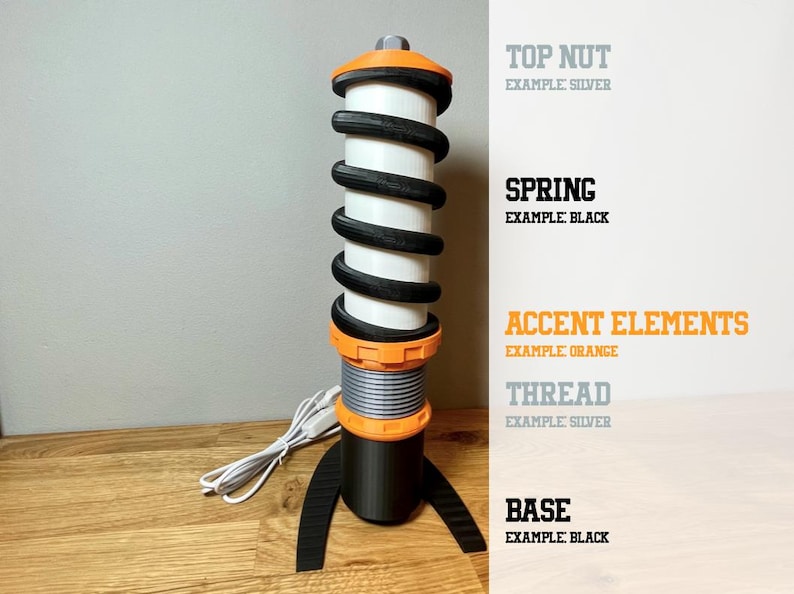Op de afbeelding: Een zwart-witte veer met oranje accenten. De veer is bevestigd aan een zwarte basis met een zilveren moer. De tekst "TOP NUT EXAMPLE SILVER", "SPRING EXAMPLE BLACK", "ACCENT ELEMENTS EXAMPLE ORANGE", "THREAD EXAMPLE SILVER" en "BASE EXAMPLE BLACK" is zichtbaar aan de rechterkant van de afbeelding.