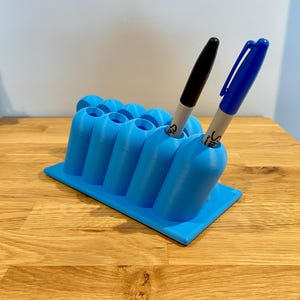 May include: A bright blue plastic desk organizer with a rectangular base and multiple cylindrical slots for pens. One black marker and one blue marker are inserted into the slots. The organizer features a grid-like arrangement of holders.