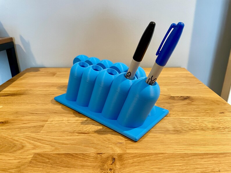 May include: A bright blue plastic desk organizer with a rectangular base and multiple cylindrical slots for pens. One black marker and one blue marker are inserted into the slots. The organizer features a grid-like arrangement of holders.