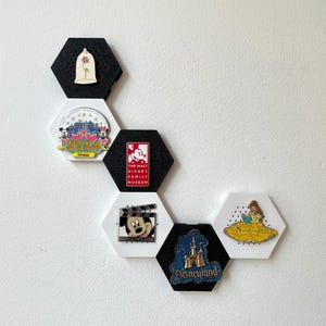 May include: A collection of Disney-themed pins displayed on black and white hexagon-shaped holders. The pins feature characters and logos from Disney films and parks, including Mickey Mouse, Belle, and Disneyland.