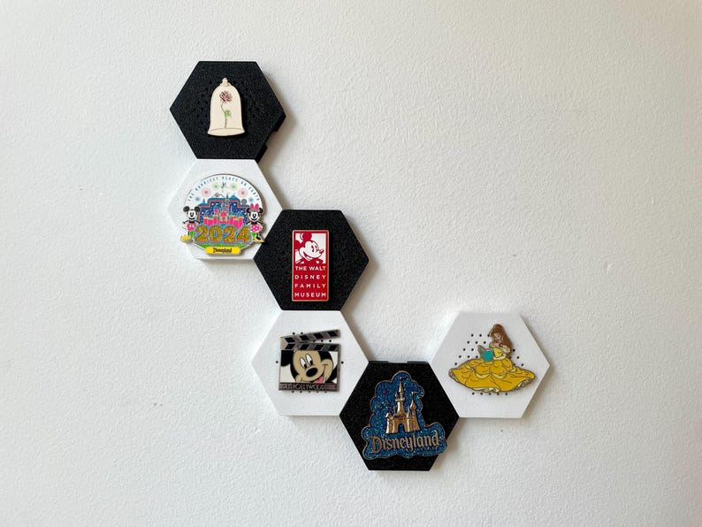 May include: A collection of Disney-themed pins displayed on black and white hexagon-shaped holders. The pins feature characters and logos from Disney films and parks, including Mickey Mouse, Belle, and Disneyland.
