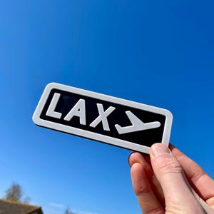 May include: White sign with black background and white lettering that reads "LAX" with a white airplane symbol pointing down.