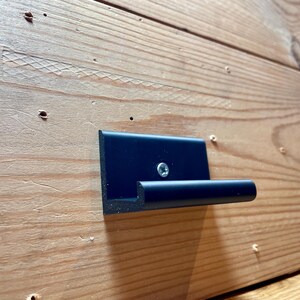 May include: A black metal hook mounted on a wooden surface. The hook has a rounded edge and a single screw.