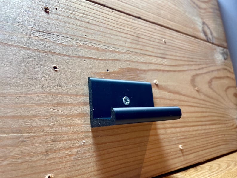 May include: A black metal hook mounted on a wooden surface. The hook has a rounded edge and a single screw.
