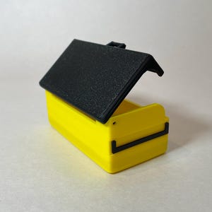 Pu&ograve; includere: Una piccola scatola gialla con un coperchio nero strutturato e incernierato. La scatola ha una forma rettangolare con dettagli neri ed &egrave; aperta, rivelando l'interno. Lo sfondo &egrave; una superficie bianca sporca.