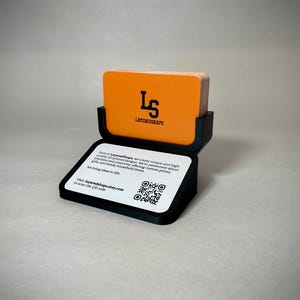 May include: A black business card holder with a stack of orange cards. The cards have the logo "LS LAYEREDSHAPE" in black. The holder has a white base with text and a QR code. The text reads "Visit layeredshape.etsy.com or scan the QR code."