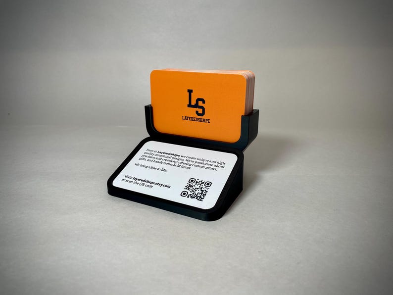May include: A black business card holder with a stack of orange cards. The cards have the logo "LS LAYEREDSHAPE" in black. The holder has a white base with text and a QR code. The text reads "Visit layeredshape.etsy.com or scan the QR code."