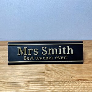 name plaque / plate with a teachers name and best teacher ever! text on the front. Black with bronze text on a wooden desk with grey background