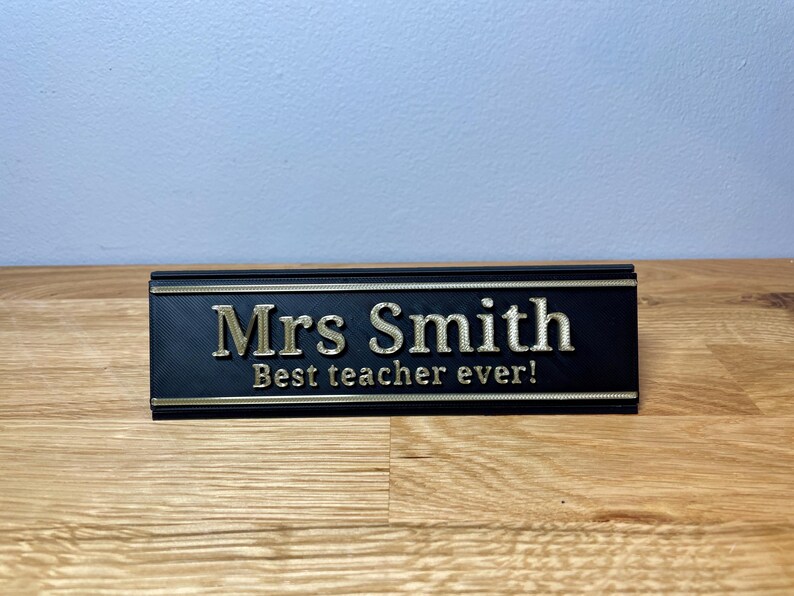 name plaque / plate with a teachers name and best teacher ever! text on the front. Black with bronze text on a wooden desk with grey background
