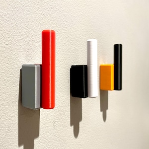 Op de afbeelding: Vier wandhaken in verschillende kleuren: rood, wit, zwart en geel. De haken zijn gemaakt van plastic en hebben een eenvoudig, modern ontwerp.