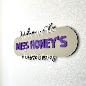 K&ouml;nnte beinhalten: Ein Klassenzimmerschild mit dem Text "Willkommen in Miss Honeys Klassenzimmer". Das Schild hat einen beigen ovalen Hintergrund mit lila Schrift und wei&szlig;em Schriftzug. Es ist an einer wei&szlig;en Wand befestigt.