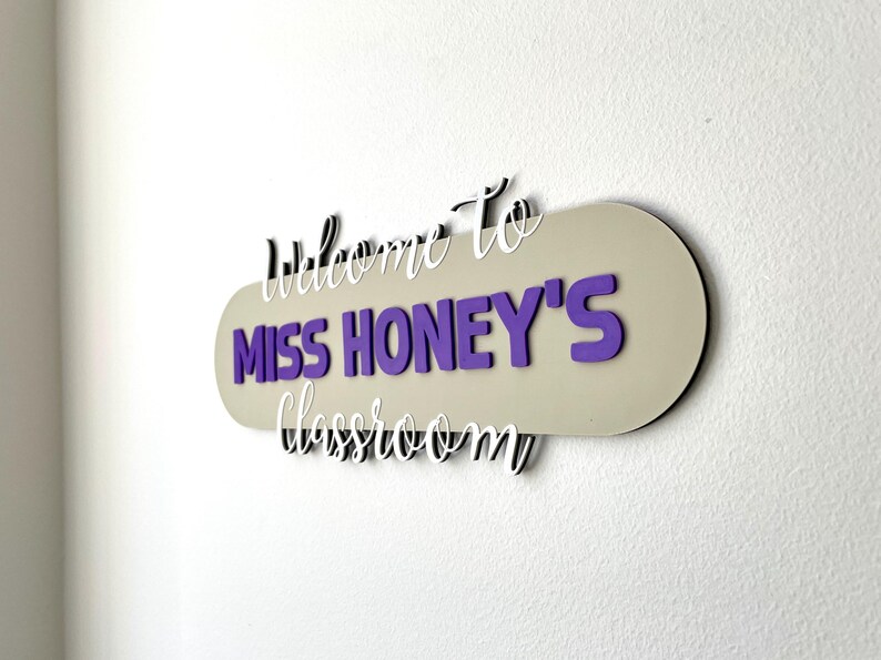 K&ouml;nnte beinhalten: Ein Klassenzimmerschild mit dem Text "Willkommen in Miss Honeys Klassenzimmer". Das Schild hat einen beigen ovalen Hintergrund mit lila Schrift und wei&szlig;em Schriftzug. Es ist an einer wei&szlig;en Wand befestigt.