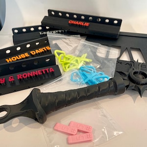 May include: An assortment of black dartboard backings with colorful text, including "CHARLIE", "HOUSE DARTS", and "RONNETTA". Also, a black sword-like object, and small plastic pieces in bags. Various colors are visible.
