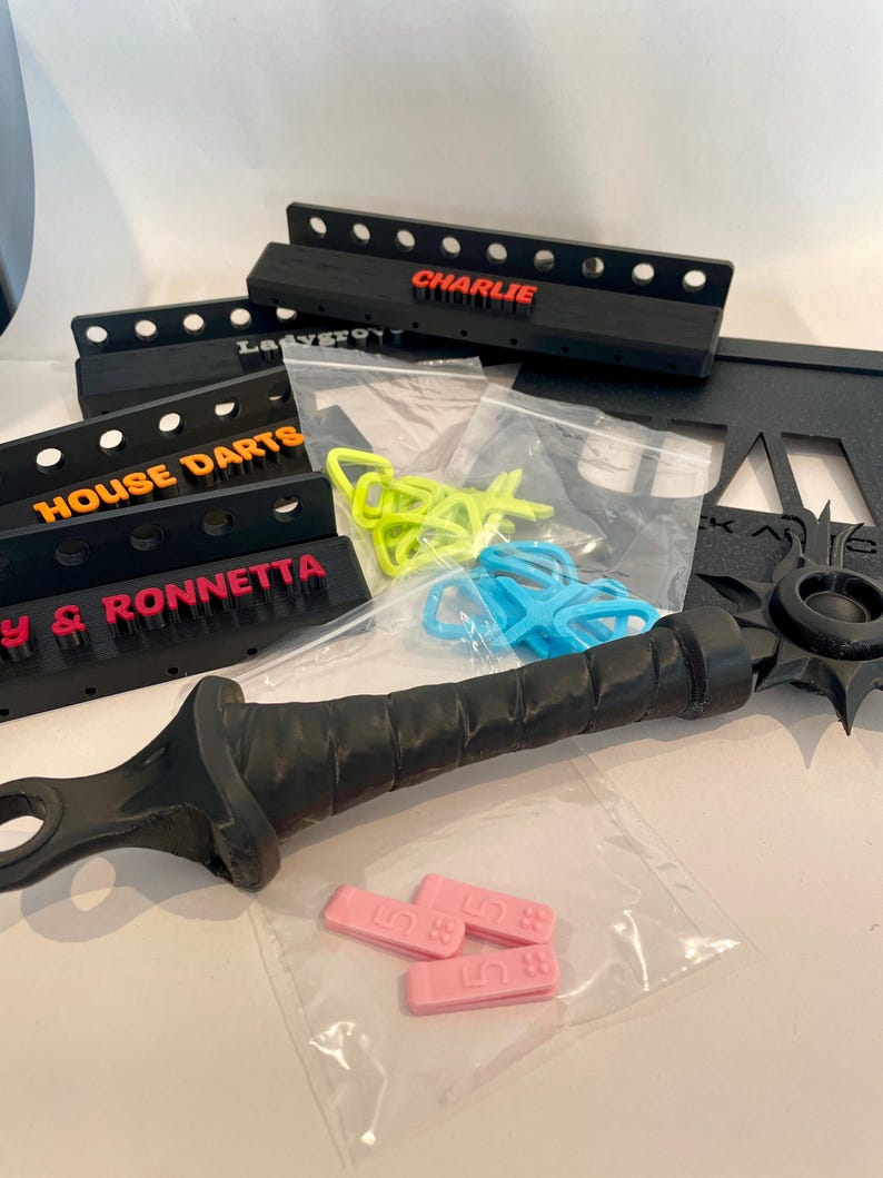 May include: An assortment of black dartboard backings with colorful text, including "CHARLIE", "HOUSE DARTS", and "RONNETTA". Also, a black sword-like object, and small plastic pieces in bags. Various colors are visible.