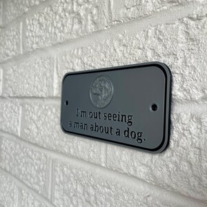 May include: A gray rectangular sign with rounded corners is mounted on a white brick wall. The sign has black trim and the text "I'm out seeing a man about a dog." A dog emblem is at the top.
