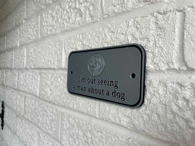 May include: A gray rectangular sign with rounded corners is mounted on a white brick wall. The sign has black trim and the text "I'm out seeing a man about a dog." A dog emblem is at the top.