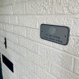 May include: A rectangular, gray metal sign with rounded corners is mounted on a white brick wall. The sign features the phrase "I'm out seeing a man about a dog." in black text. A circular emblem is at the top of the sign.