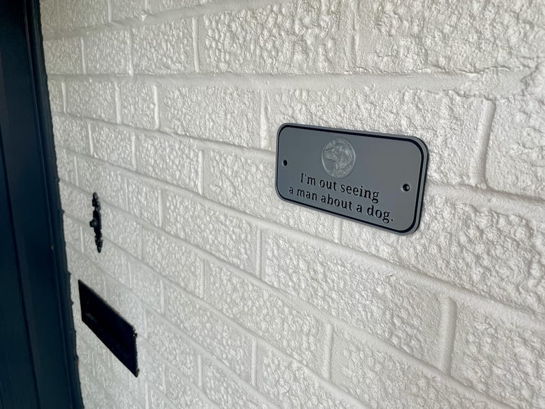May include: A rectangular, gray metal sign with rounded corners is mounted on a white brick wall. The sign features the phrase "I'm out seeing a man about a dog." in black text. A circular emblem is at the top of the sign.