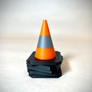 May include: A bright orange and grey traffic cone sits on a stack of black geometric coasters. The cone has a bright orange top and bottom with a grey band in the middle. The coasters are stacked in a staggered pattern.