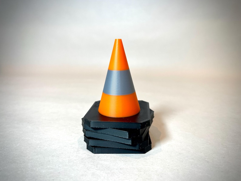 May include: A bright orange and grey traffic cone sits on a stack of black geometric coasters. The cone has a bright orange top and bottom with a grey band in the middle. The coasters are stacked in a staggered pattern.