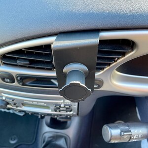 May include: A black car vent phone mount with a knob to adjust the angle. The mount is attached to a car's air vent.