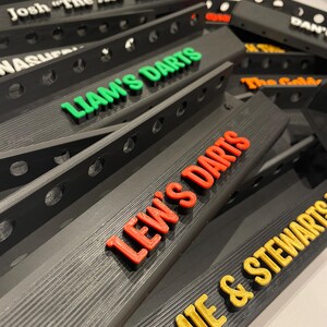 May include: A collection of black dartboard backboards with various names in colorful 3D lettering. Names include "Josh 'The Ace' Allen", "Liam's Darts", "Lew's Darts", and "Jamie & Stewarts". The backboards have a series of holes along the top edge.