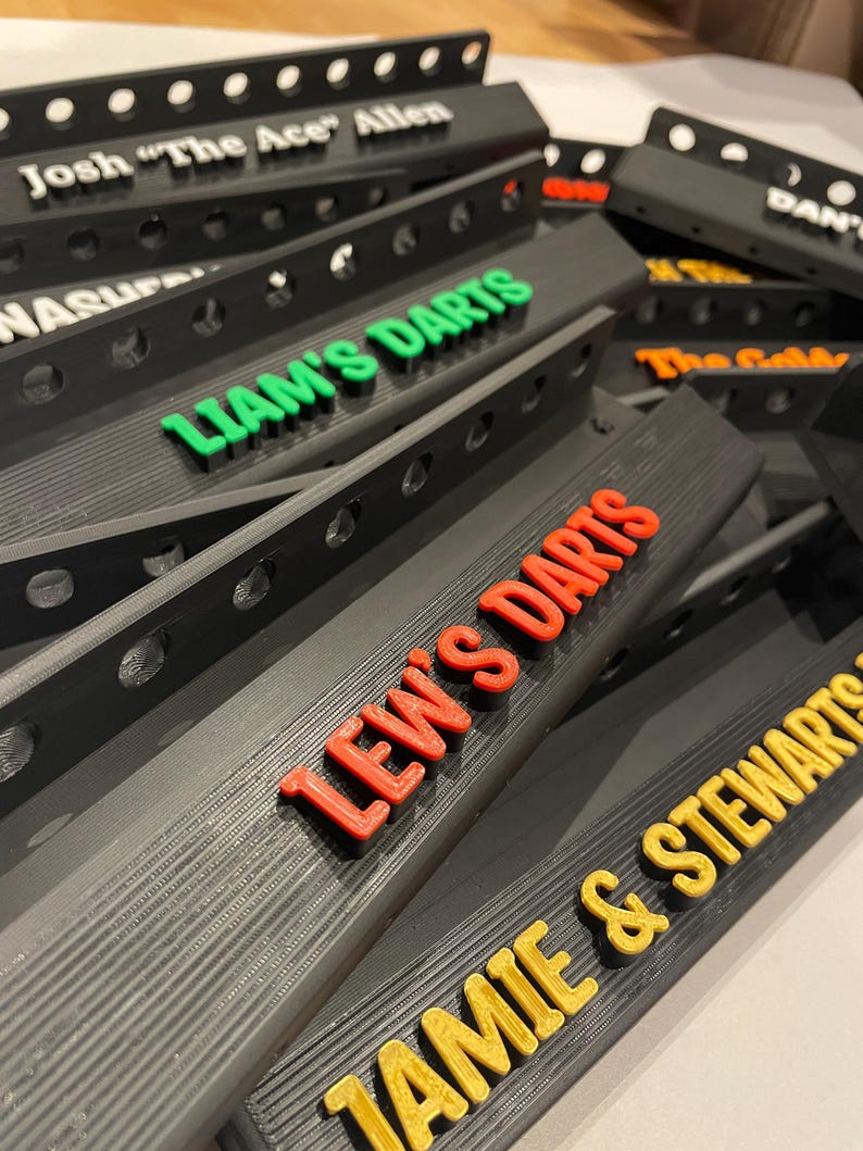 May include: A collection of black dartboard backboards with various names in colorful 3D lettering. Names include "Josh 'The Ace' Allen", "Liam's Darts", "Lew's Darts", and "Jamie & Stewarts". The backboards have a series of holes along the top edge.