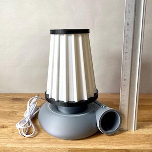 Puede incluir: Una l&aacute;mpara de mesa gris y blanca con una pantalla de tela plisada y una base redonda. La l&aacute;mpara tiene un cable y un enchufe y un borde negro alrededor de la pantalla.