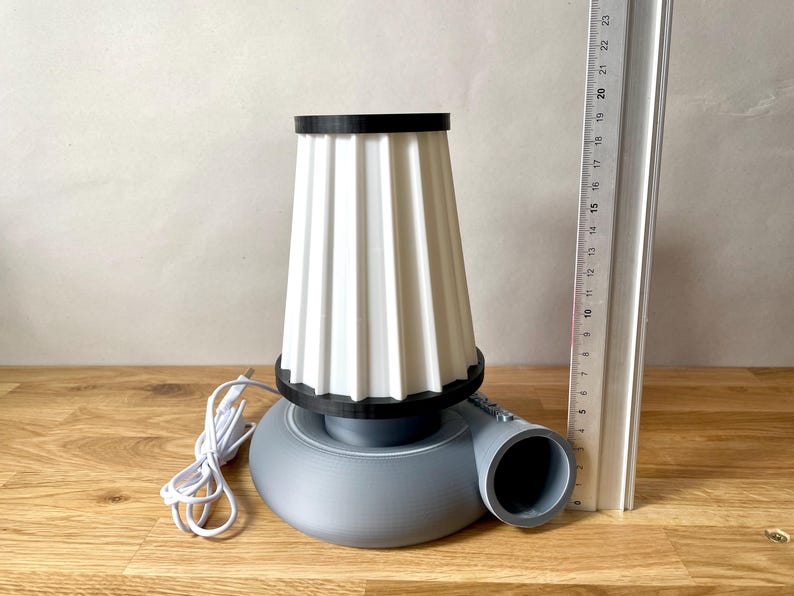 Puede incluir: Una l&aacute;mpara de mesa gris y blanca con una pantalla de tela plisada y una base redonda. La l&aacute;mpara tiene un cable y un enchufe y un borde negro alrededor de la pantalla.