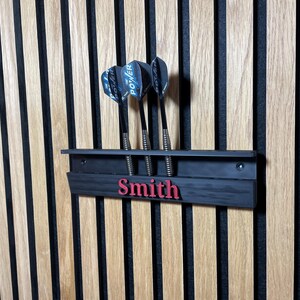 May include: A black dart holder with three darts, mounted on a wooden wall. The holder has the name "Smith" in red lettering. The darts have black and blue flights.