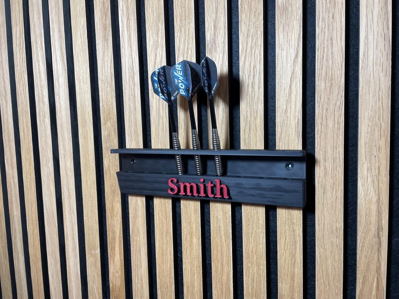 May include: A black dart holder with three darts, mounted on a wooden wall. The holder has the name "Smith" in red lettering. The darts have black and blue flights.
