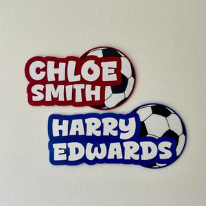 May include: Two personalized soccer-themed name tags. One is red with the name "CHLOE SMITH" and a soccer ball graphic. The other is blue with the name "HARRY EDWARDS" and a soccer ball graphic.