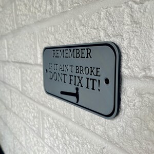 May include: A rectangular gray metal sign with black text that reads "REMEMBER IF IT AIN'T BROKE DON'T FIX IT!" A black hammer graphic is at the bottom. The sign is mounted on a white brick wall.